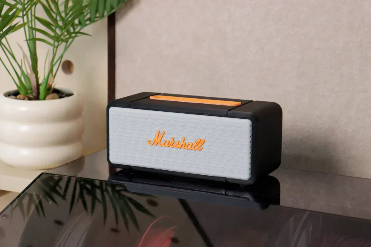 Ein 3D-gedrucktes Miniatur-Lautsprechermodell mit grauem Netz und orangefarbenem Logo steht neben einem Blumentopf auf dem Tisch.