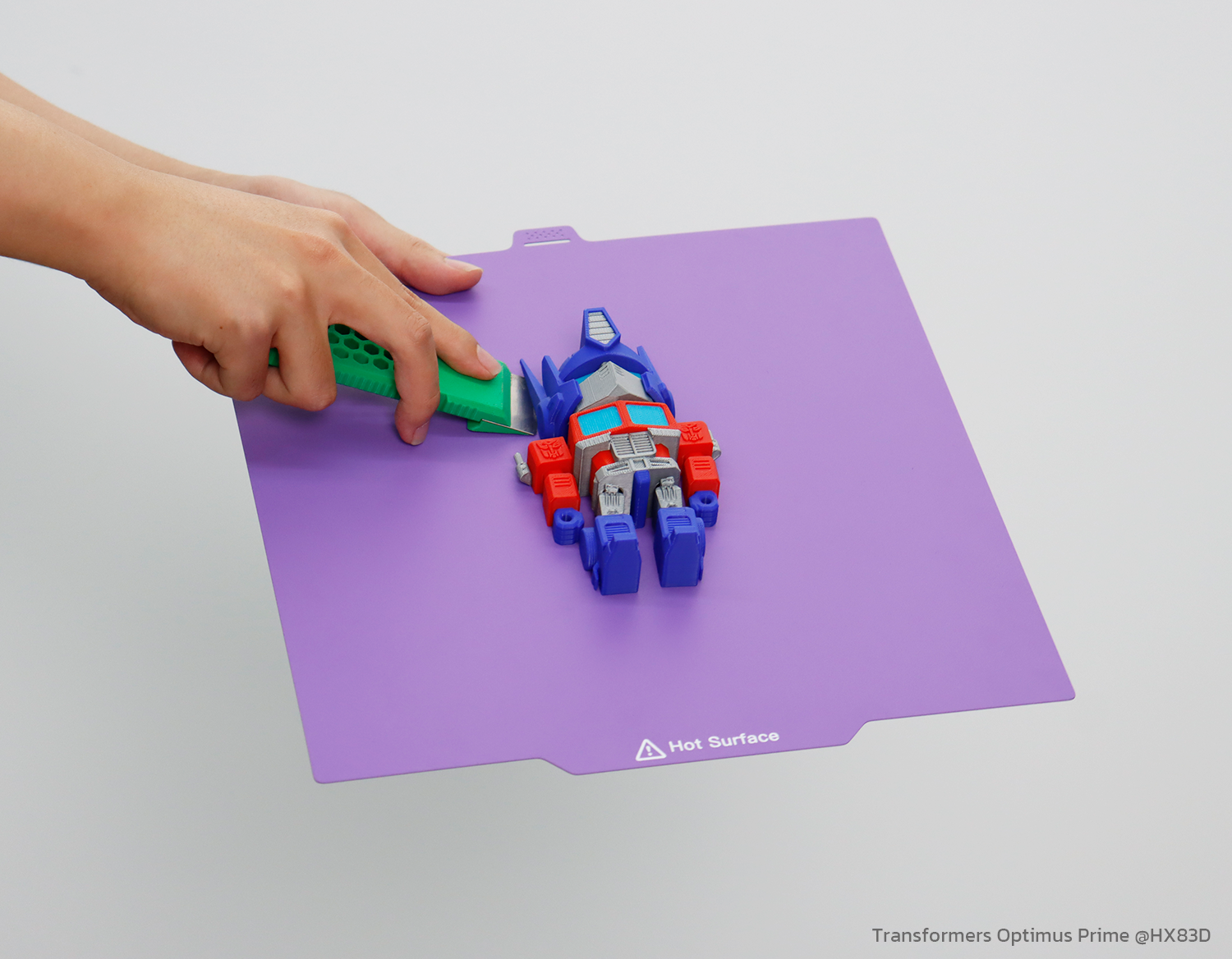 A hand lifts a red, white, and blue Optimus Prime-style toy from a purple 3D printer build plate using a green tool